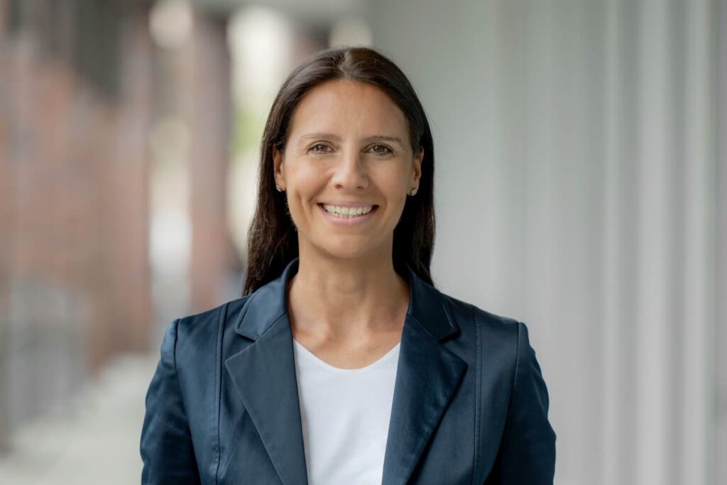 Porträt einer Frau mit dunklem Haar im dunkelblauen Blazer und weißem Oberteil vor hellem Hintergrund.