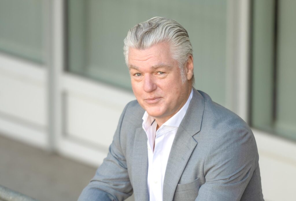 Porträt eines Geschäftsmanns mit grauem Haar im hellgrauen Anzug, sitzend vor moderner Gebäudefassade.
