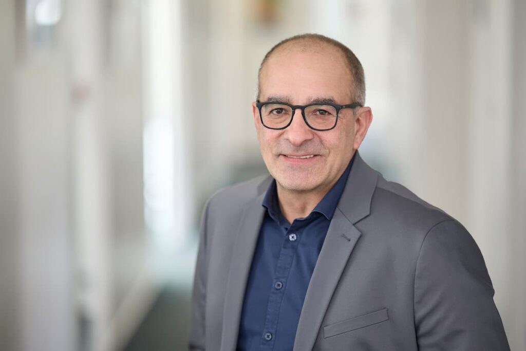 Porträt eines Mannes mit Brille im grauen Sakko und dunkelblauem Hemd vor hellem Hintergrund.